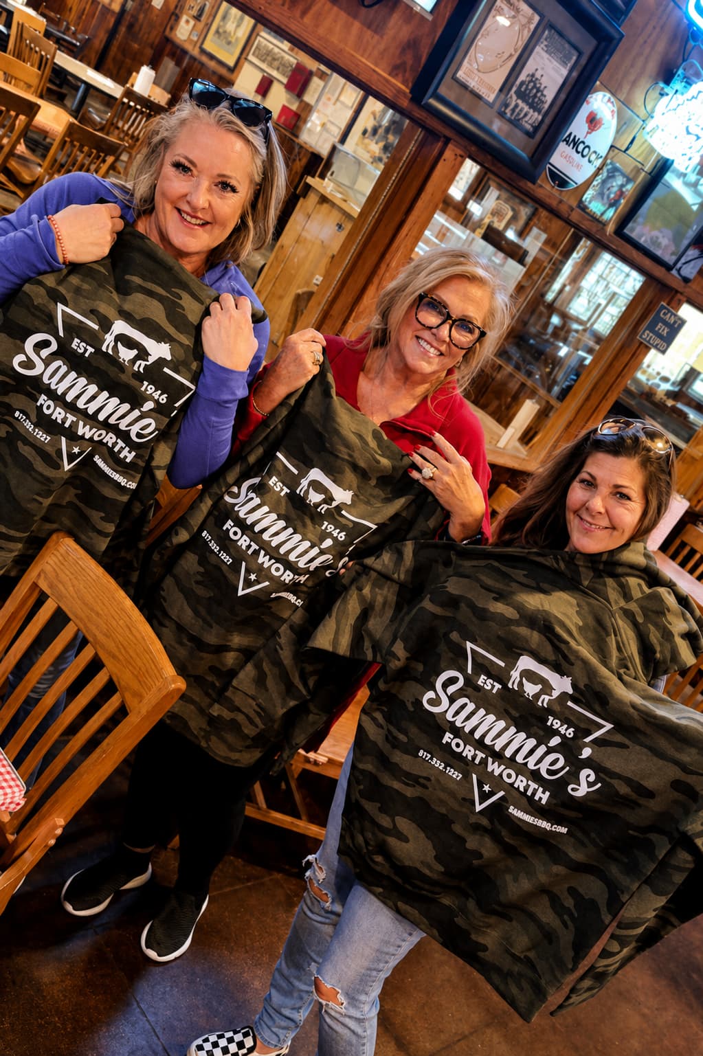Three friends holding up camo Sammie's hoodies in the dining room