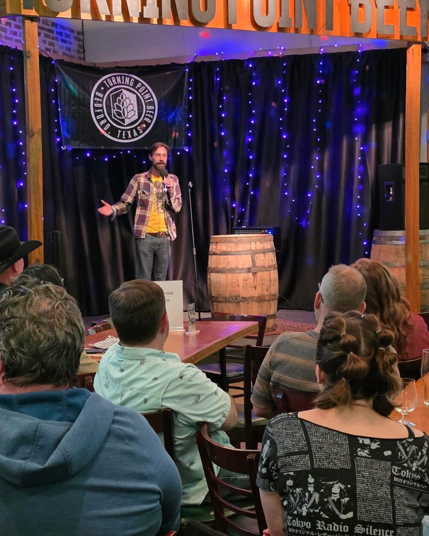 A comedian on the taproom stage under string lights with the crowd seated at tables