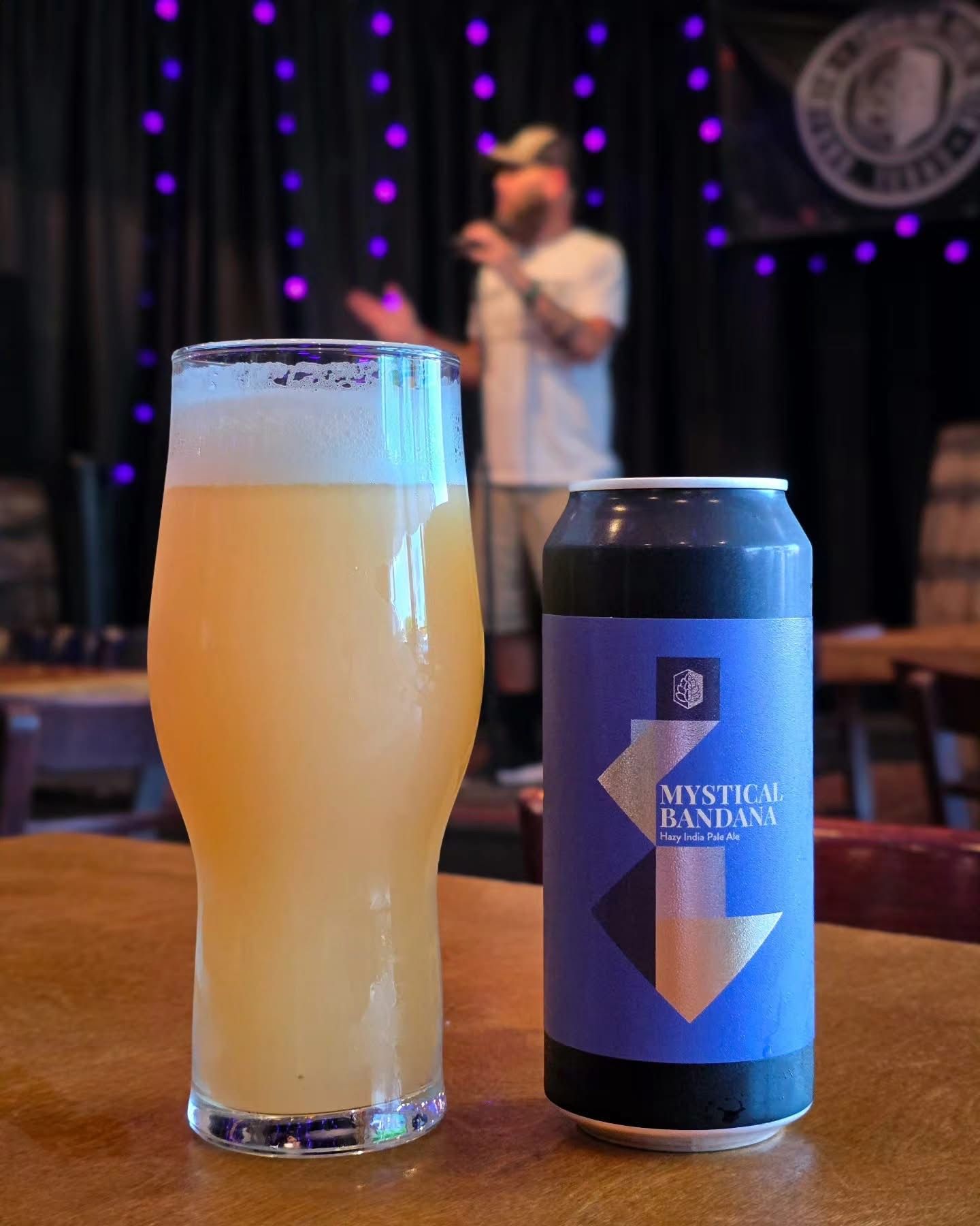 A hazy pour and a Mystical Bandana can on a table with a performer on stage behind