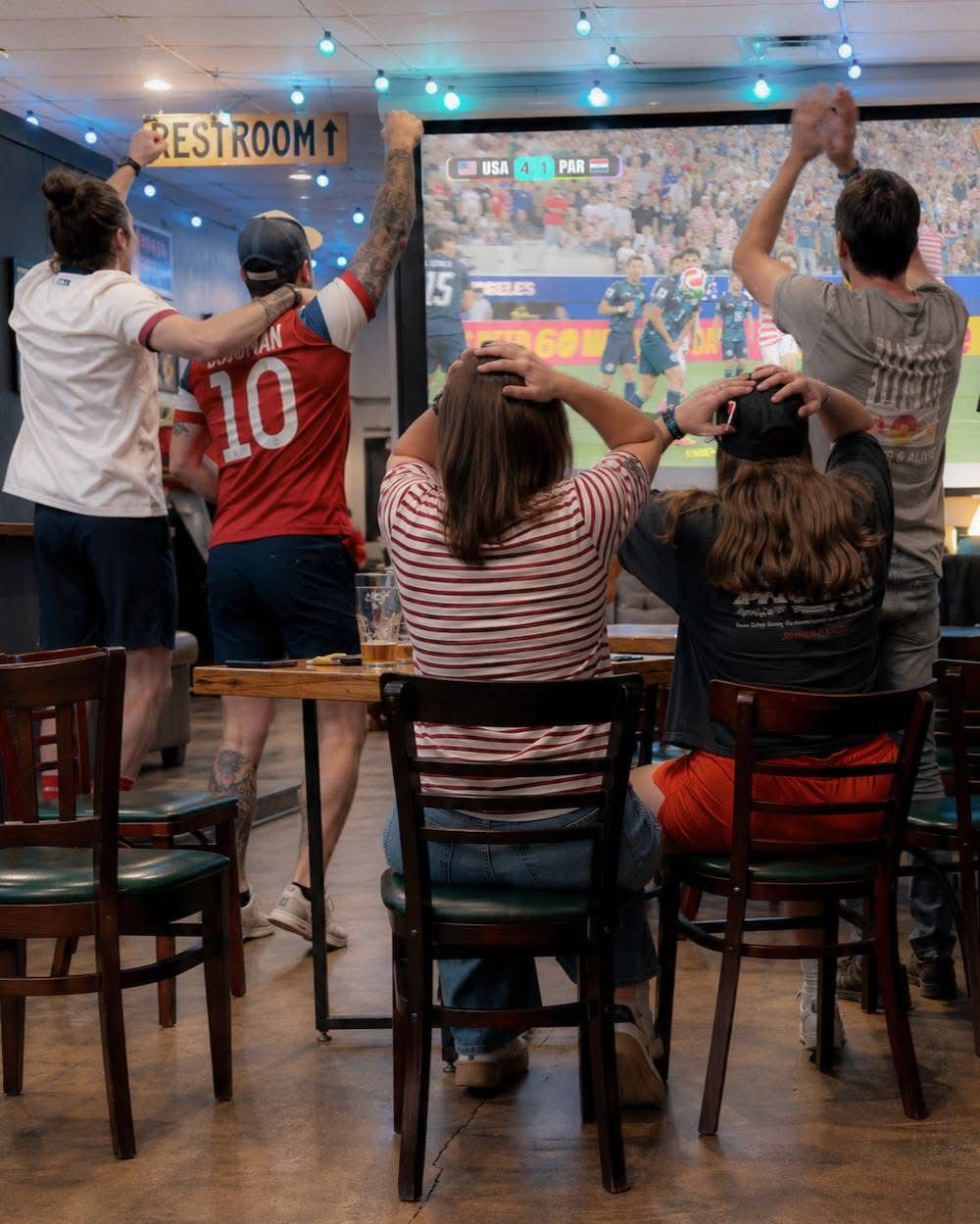 Fans at high-top tables cheering at a soccer match on the projector screen
