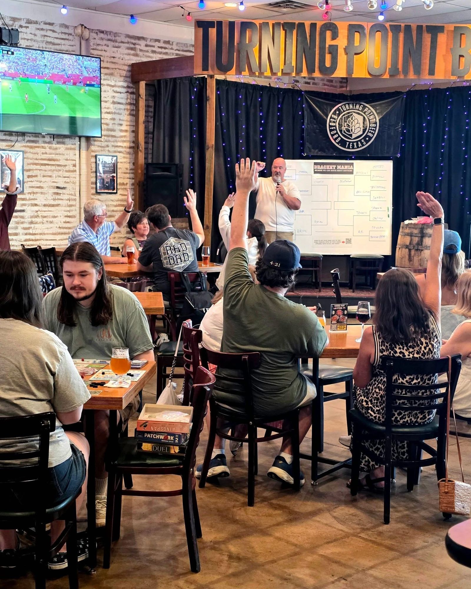 The trivia host beside a giant bracket board as players raise their hands at the tables
