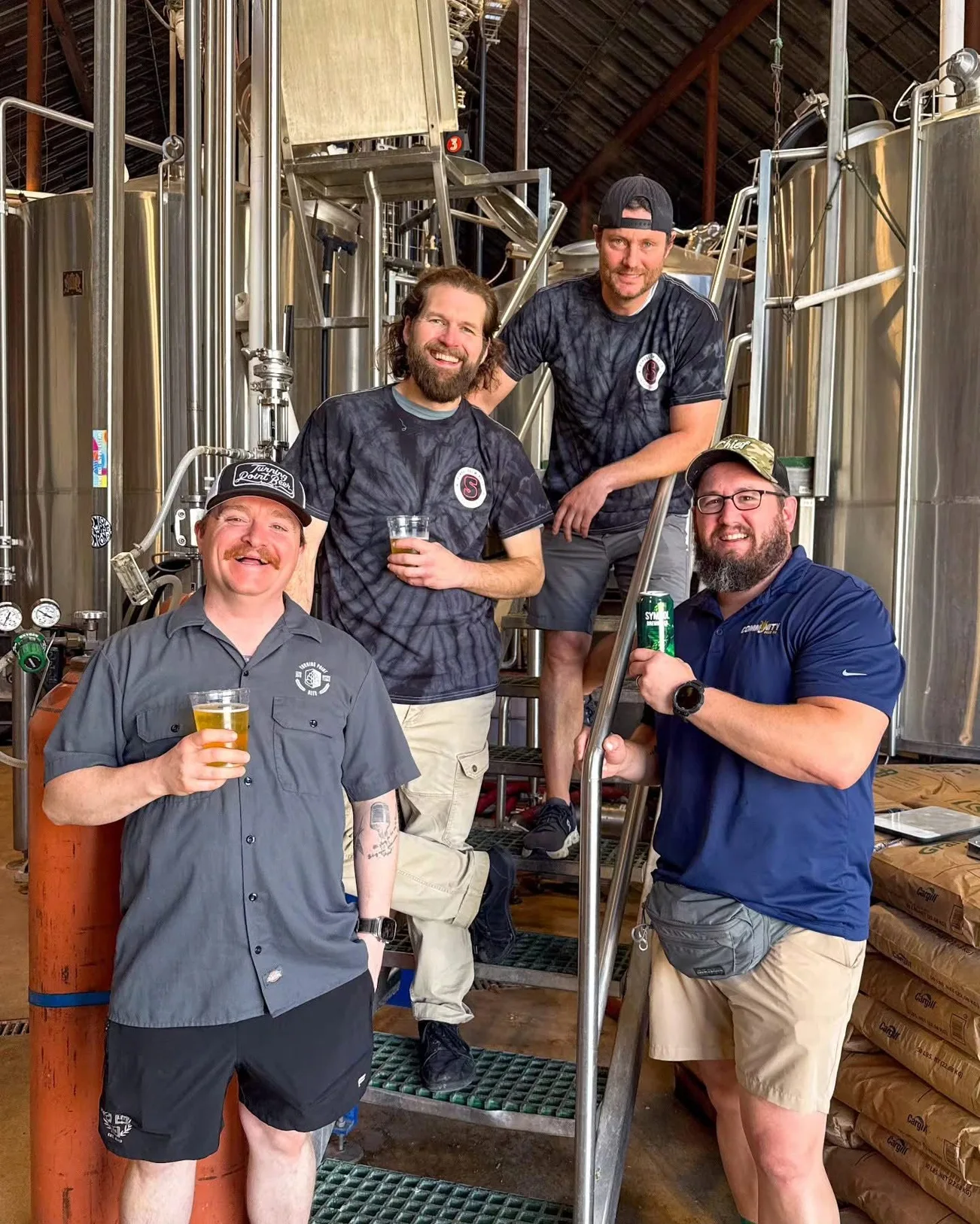 The four-person brew team with pours and cans in hand on the brewhouse stairs between stainless tanks