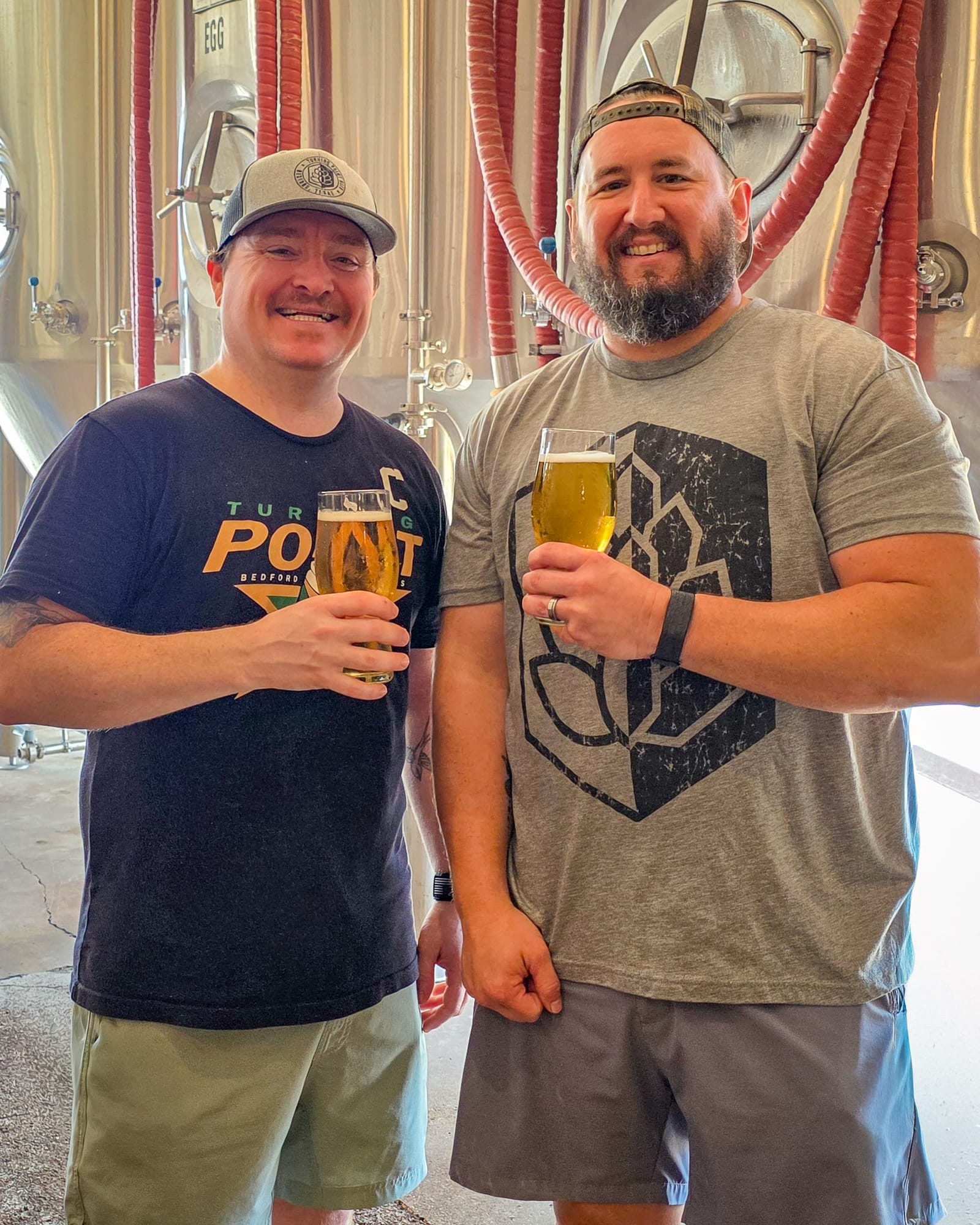 Two Turning Point brewers in branded tees raising glasses of pale beer in front of the stainless fermenters